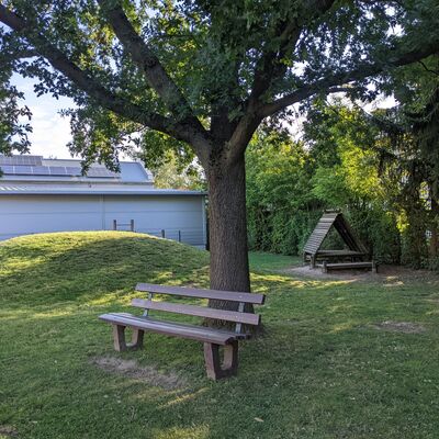 Bild vergrößern: Außengelände mit Bank vor einem Baum. Im Hindergrund eine kleine Holzhütte zum Spielen.